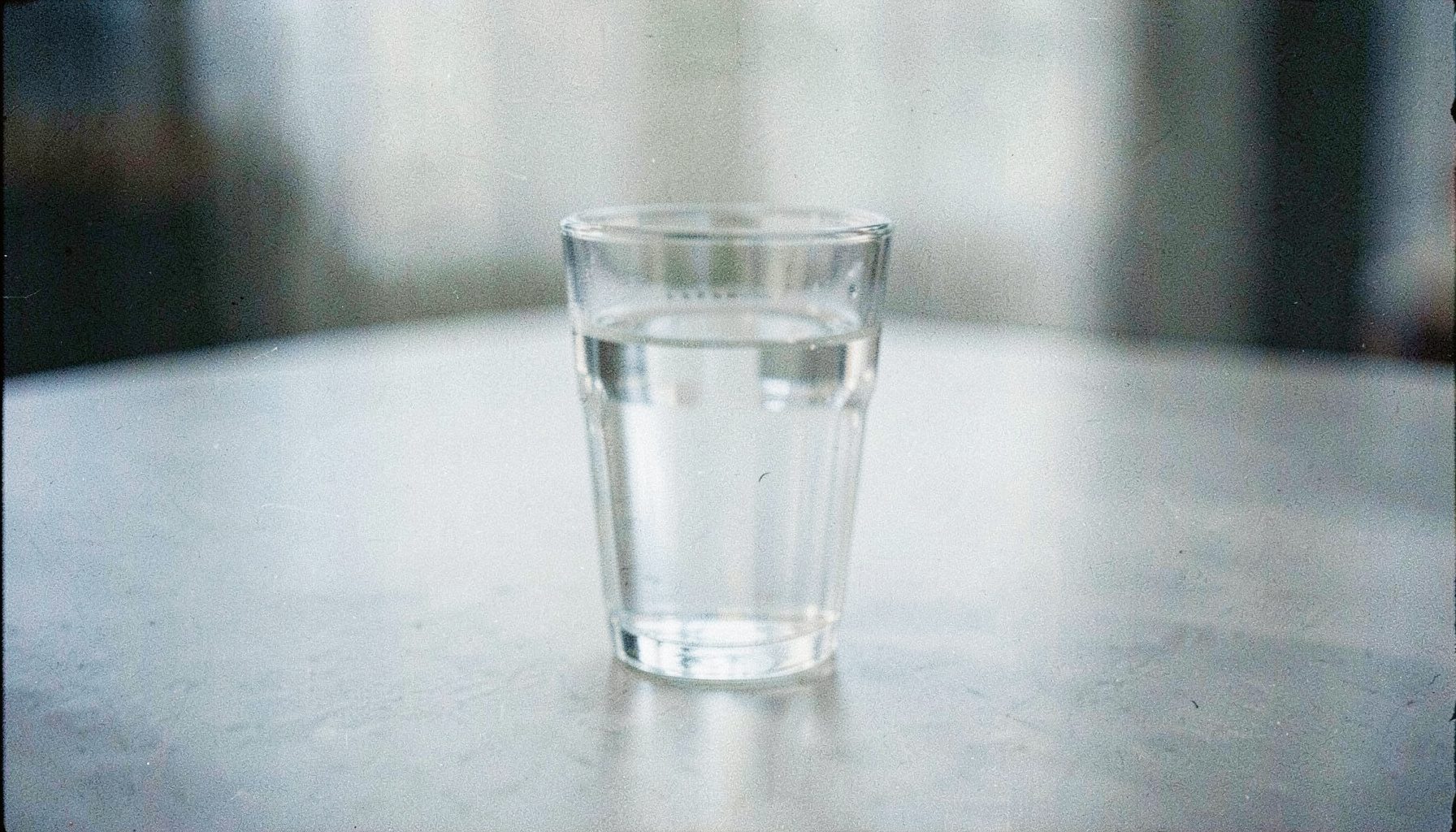 A grainy analog film photograph of a glass of water on a plain tabletop, soft natural light, muted grays and pale blues, contemplative mood.